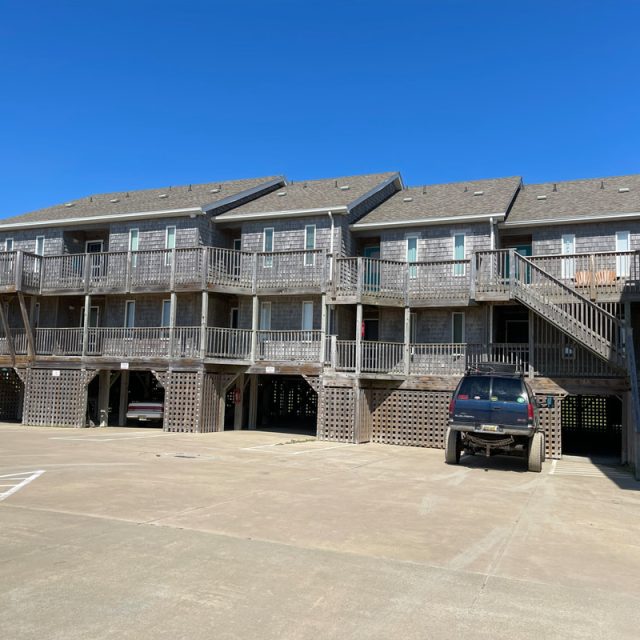 Two-story wooden apartment building on stilts with exterior stairs and parked vehicles underneath.