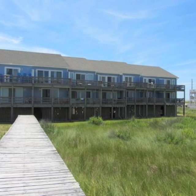 A two-story blue beach house with wide decks, viewed from a wooden boardwalk across grassy dunes under a blue sky.