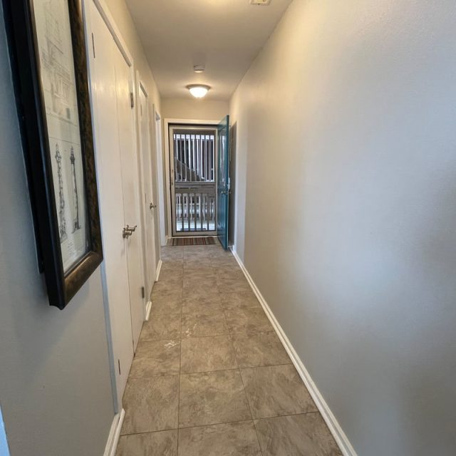 A narrow hallway with tile floor, white walls, doors on the left, and an open door leading outside at the end.