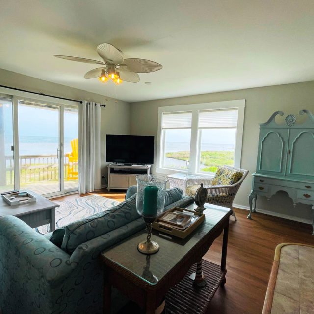 Bright living room with large windows, a TV, blue furniture, and a view of a balcony overlooking water and greenery.