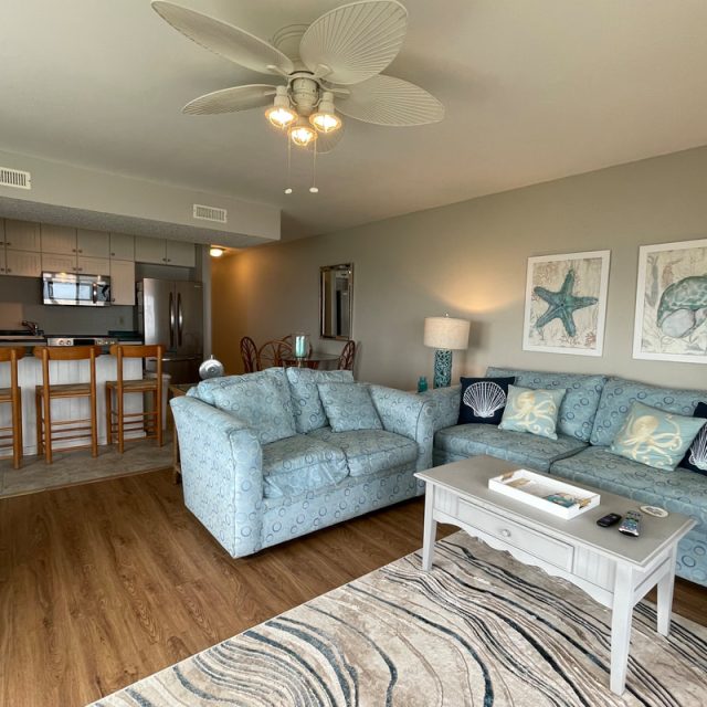 Living room with blue couches, a coffee table, and beach-themed decor, adjacent to a kitchen with barstools.