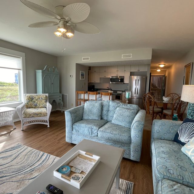 Cozy beach-themed living room and kitchen with light blue furniture, wicker chairs, and a view of greenery.