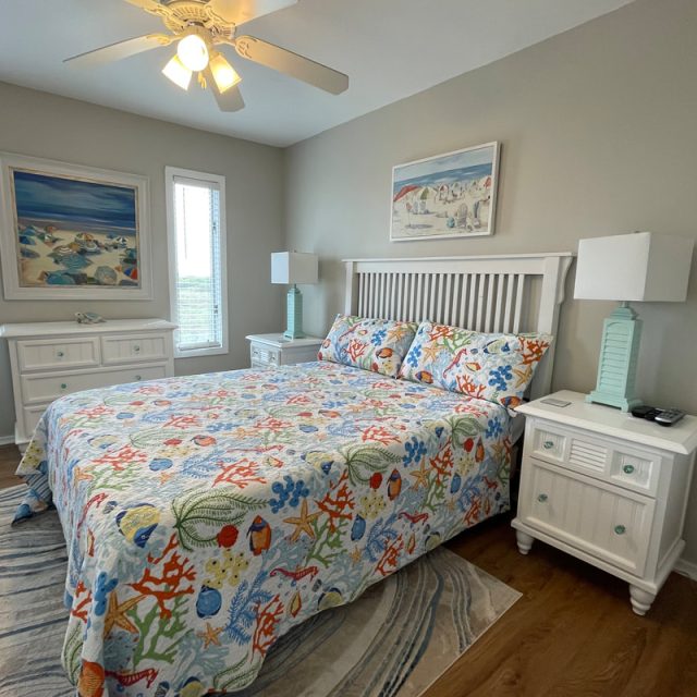 Bedroom with a king bed, coastal-themed bedding, two nightstands, two lamps, and seaside wall art.