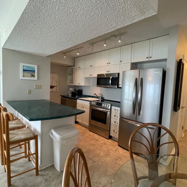 Modern kitchen with white cabinets, stainless steel appliances, green countertop, and a small dining area with four chairs.