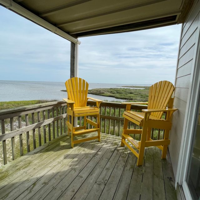 Two yellow chairs on a wooden porch overlook grassy wetlands and a body of water under a cloudy sky.