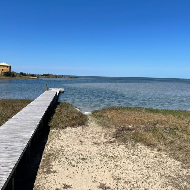 Wooden dock leads to calm water with grassy shore; a yellow house sits in the distance under a clear blue sky.