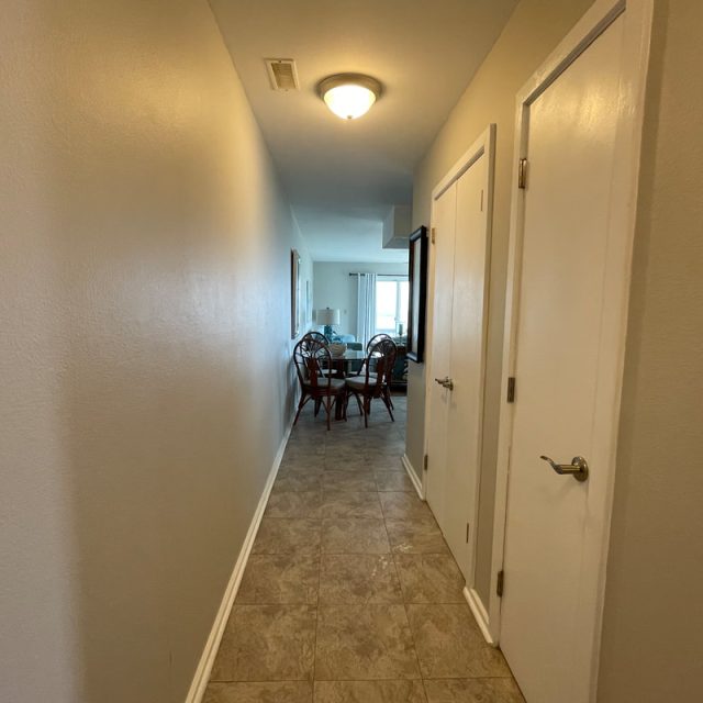 A narrow hallway with tiled floor leads to a dining area with a table and chairs, lit by a ceiling light.