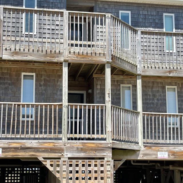 Two-story wooden beach house with weathered shingles, large porches, and white framed windows under a sunny sky.