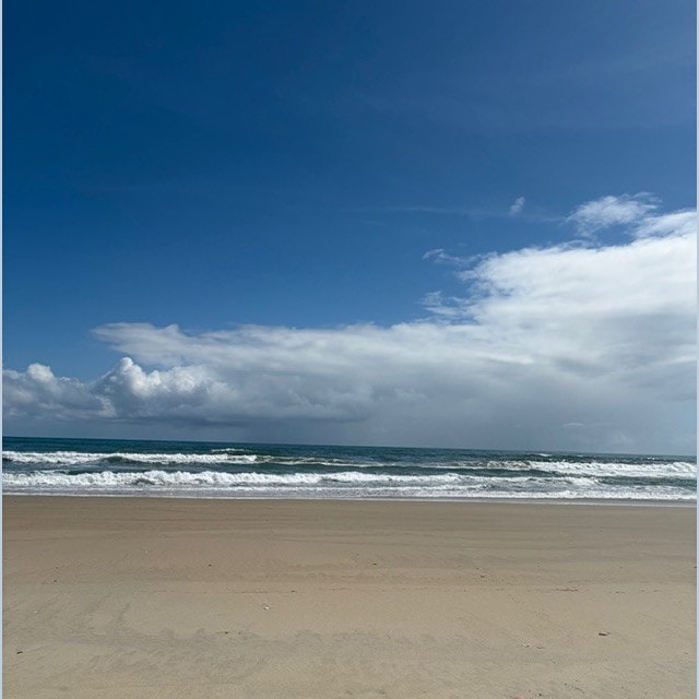 Sandy beach with gentle waves under a clear blue sky and scattered clouds.