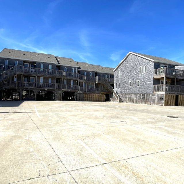 Two gray wooden buildings with multiple decks stand beside an empty concrete parking lot under a clear blue sky.