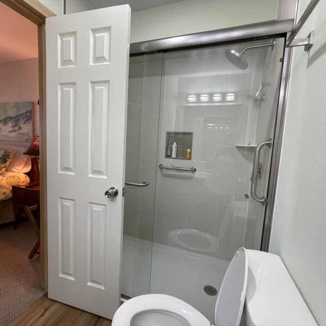 Open bathroom door reveals a glass shower, white toilet, and glimpse of a bedroom with floral bedding in background.