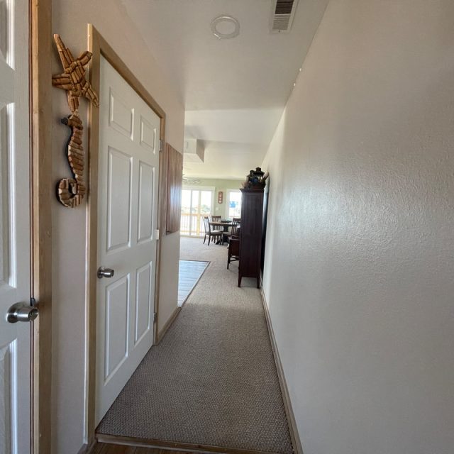 Hallway with a beige carpet, white doors, wall decor, leading to a dining area with natural light in the background.