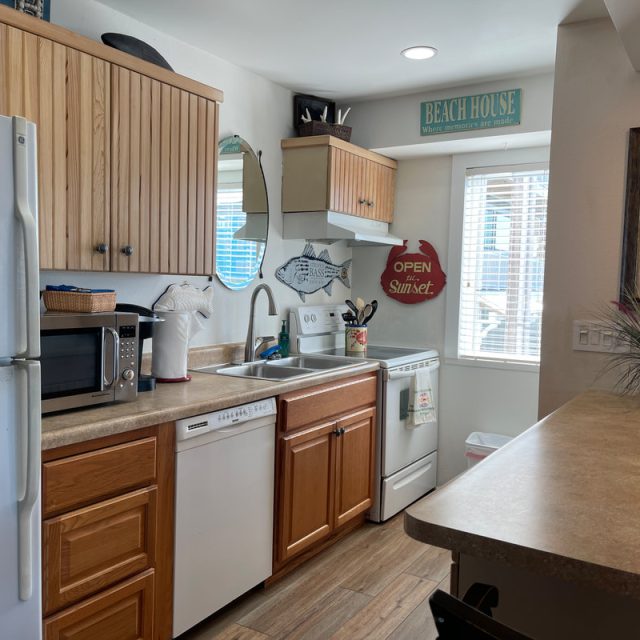 Small kitchen with white appliances, wood cabinets, beach decor, and sunlight coming through a window above the sink.