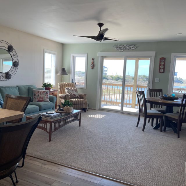 Bright living and dining area with large windows, light furniture, and a balcony overlooking water and trees.