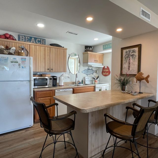 Small beach-themed kitchen with wooden cabinets, white appliances, bar counter, and three chairs.