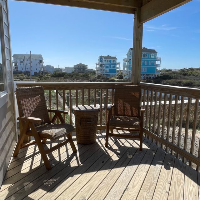 Wooden deck with two wicker chairs and a table, overlooking houses and dunes under a clear sky.