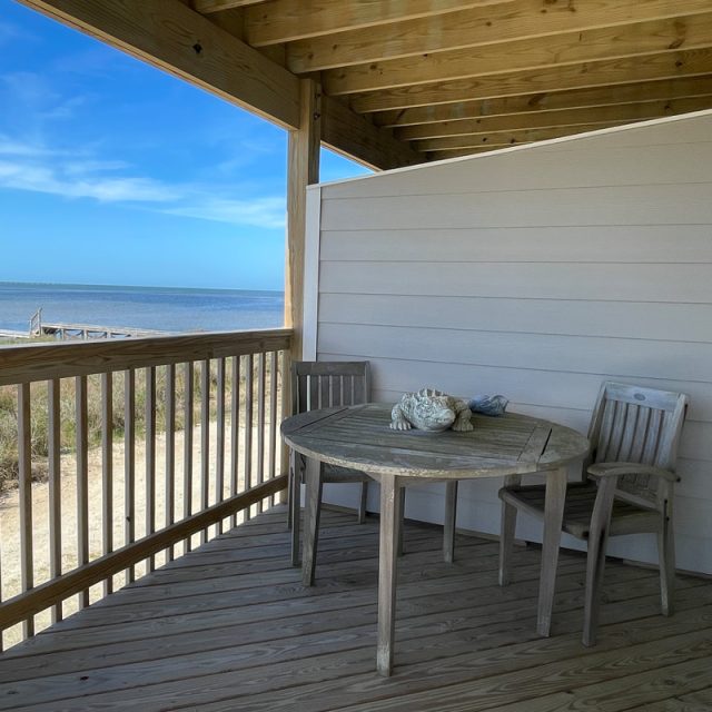 Wooden deck with two chairs and a round table overlooks the beach and ocean under a clear blue sky.