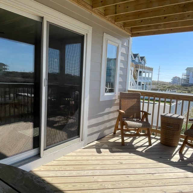 Wood deck with two wicker chairs, a small table, sliding glass door, and view of neighboring houses under clear sky.