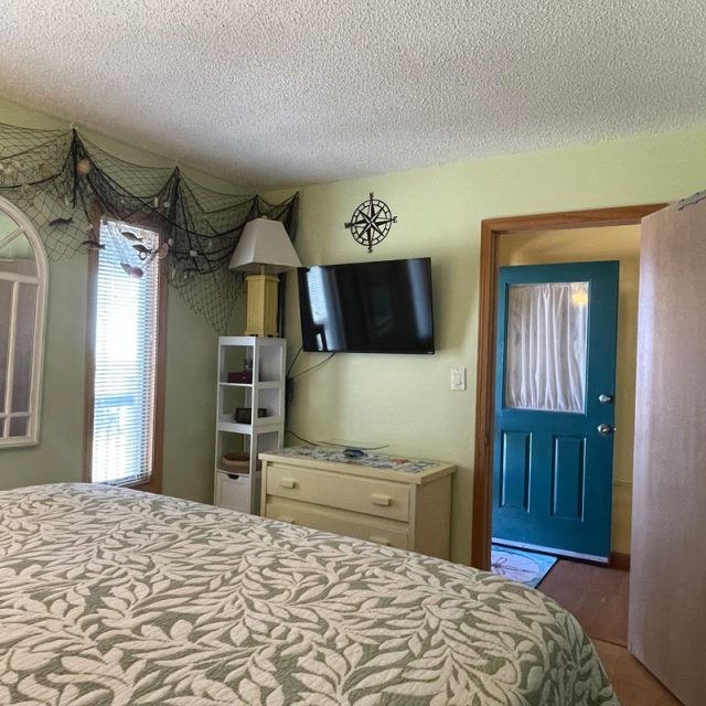 Bedroom with patterned bedspread, wall-mounted TV, dresser, open door, and decorative fishnet on light green walls.