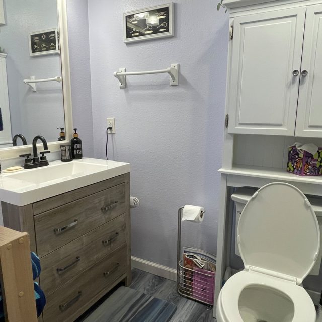 Bathroom with open toilet, vanity sink, cabinet, towels, wall shelf, and a trash can with magazines.
