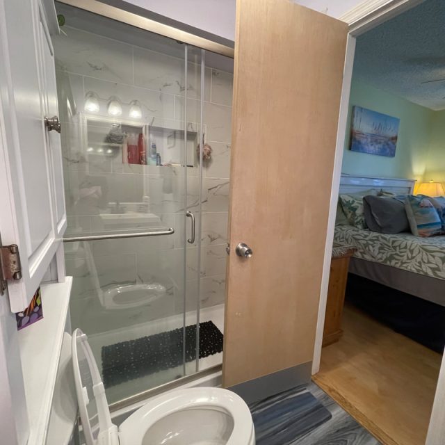 Open bathroom door reveals a toilet, shower with glass doors, and a view into a bedroom with a bed and lamp.