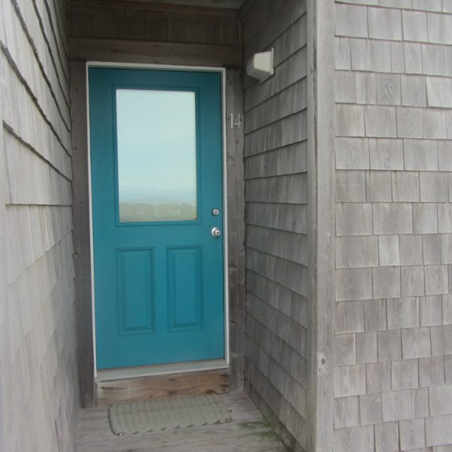 A teal door with a window, labeled 14, set in a shingle-style house with a doormat in front.