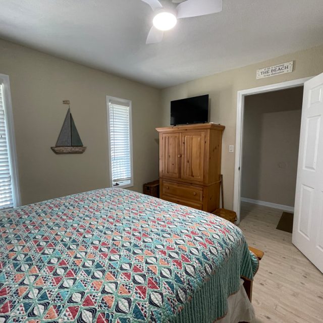 Bedroom with patterned quilt, wooden dresser with TV, two windows, and a small sailboat wall decoration.