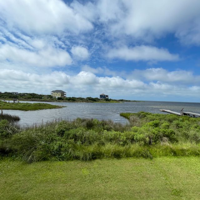 Grassy shoreline with docks, calm water, and houses in the distance under a partly cloudy blue sky.