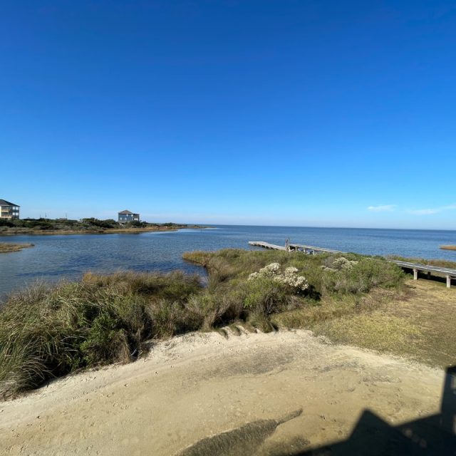 Sandy shore with grassy dunes, a wooden walkway, and two houses near a calm body of water under a clear blue sky.