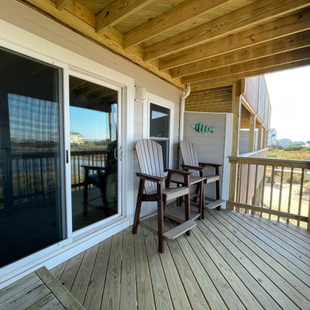 Covered wooden porch with two chairs, a small table, and a wall with fish decor; view of sand dunes outside.