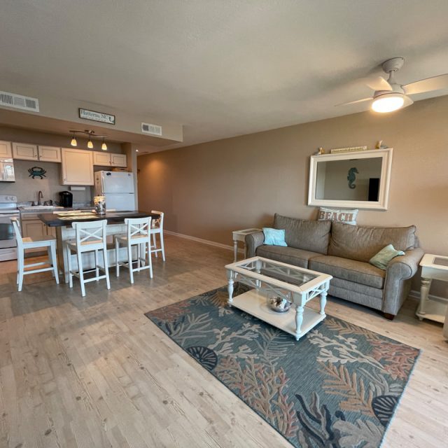 Open-plan living room and kitchen with neutral tones, beach-themed decor, and wooden flooring.