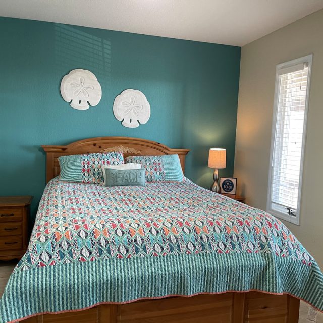 Bedroom with a wooden bed, colorful quilt, bedside tables, lamp, and beach-themed decor on teal and tan walls.
