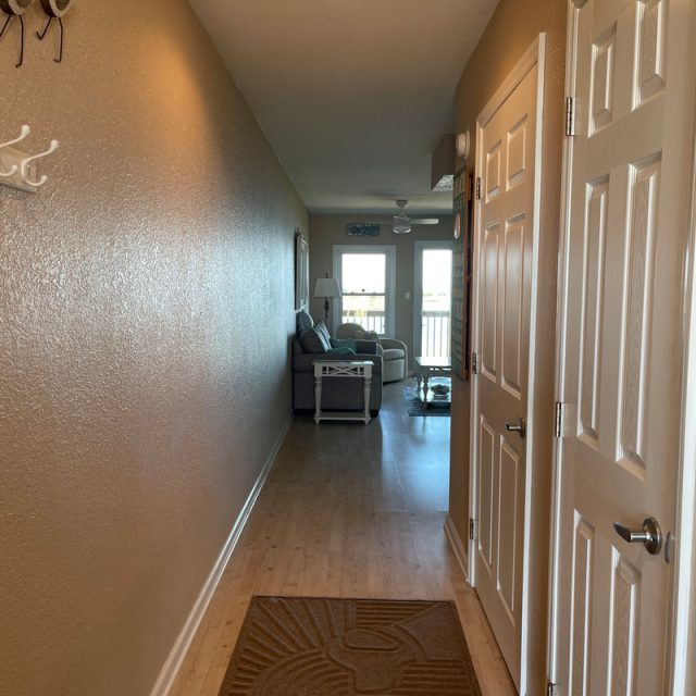 View down a hallway with tan walls, wood floor, rug, and an open door leading to a living room with windows.