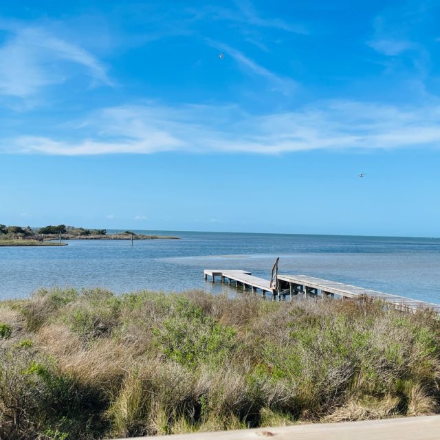 A wooden dock extends over calm coastal water, with grassy marsh in the foreground and a blue sky above.