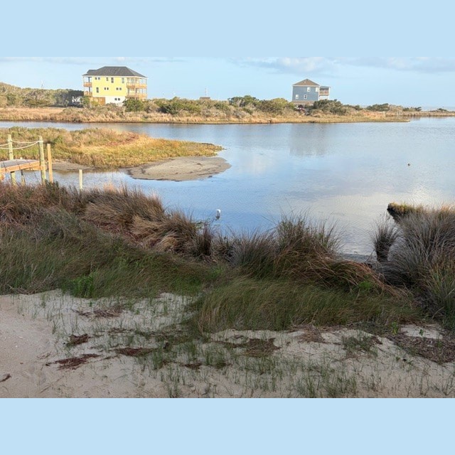 A pond bordered by tall grass with two houses in the background under a partly cloudy sky.