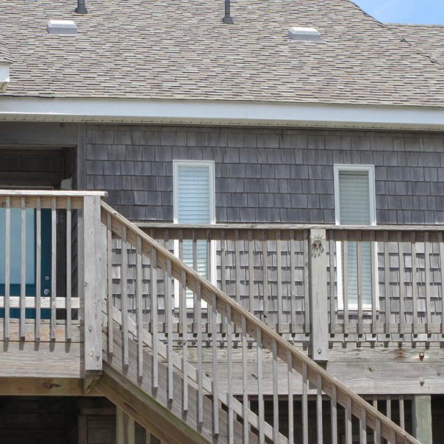 Wooden stairs and railing lead up to a beach house with gray shingles and two windows with closed blinds.