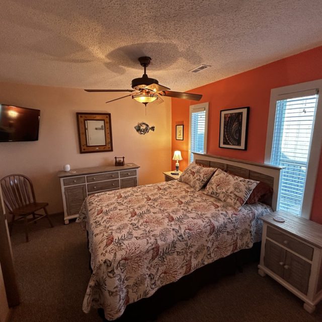 Bedroom with a double bed, two nightstands, a dresser, wall-mounted TV, chair, and two windows with blinds.