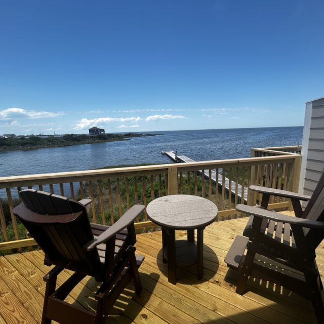 Two wooden chairs and a table on a deck overlook a calm body of water under a clear, sunny sky.