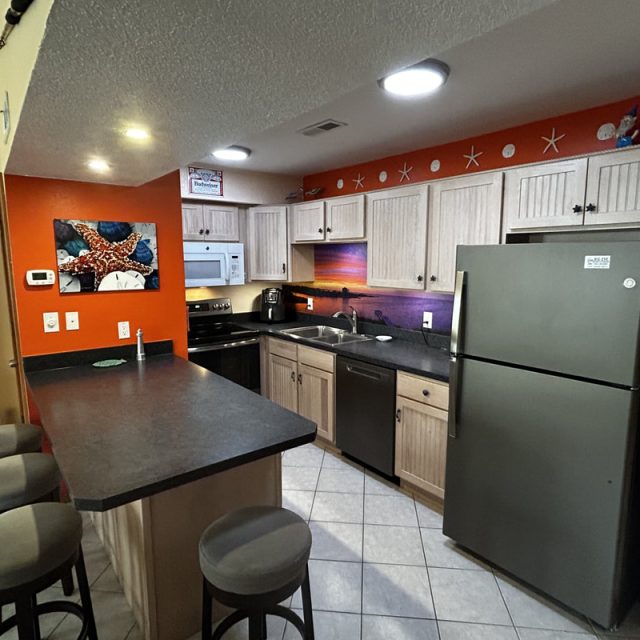 Kitchen with light wood cabinets, stainless appliances, bar counter with stools, and beach-themed wall decor.