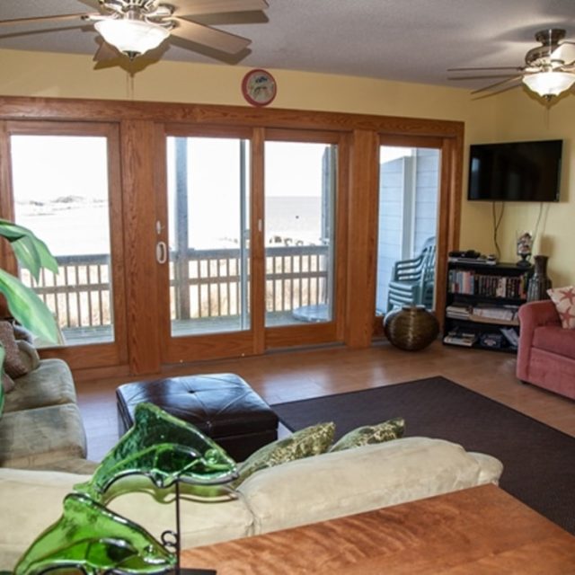 Living room with two sofas, a wall-mounted TV, ceiling fans, and glass doors opening to a patio with outdoor chairs.