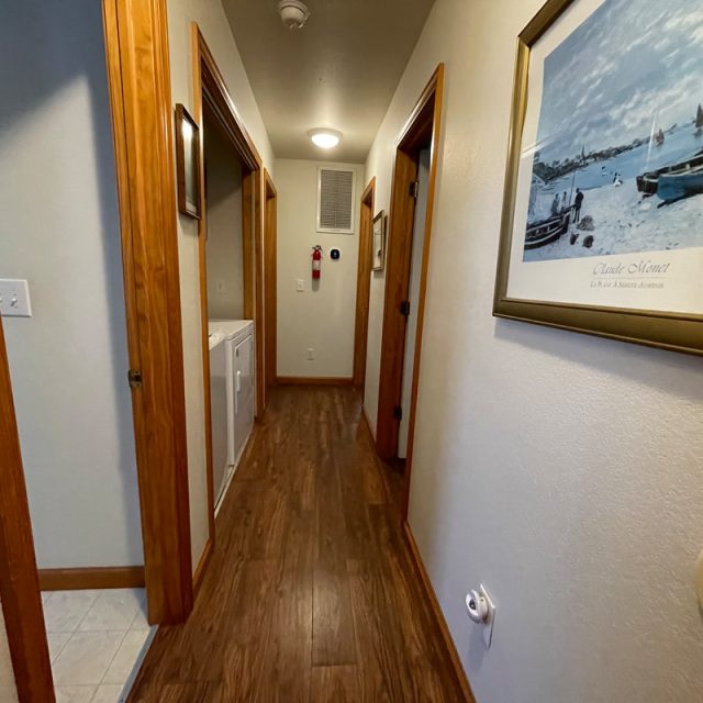 A hallway with wooden floors, open doorways, a laundry area, and a framed boating picture on the wall.