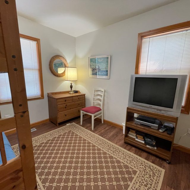 Small room with a bunk bed, dresser, chair, lamp, TV on a stand, patterned rug, and two windows with blinds.