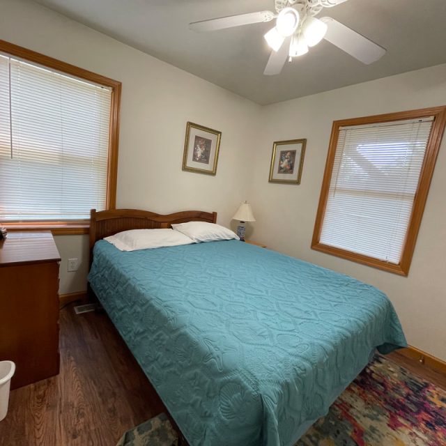Small bedroom with a double bed, teal bedspread, wooden furniture, two windows, and three framed pictures on the wall.