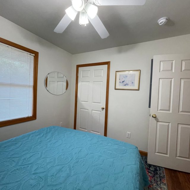 Small bedroom with a blue bedspread, two white doors, a round mirror, and a window with closed blinds.
