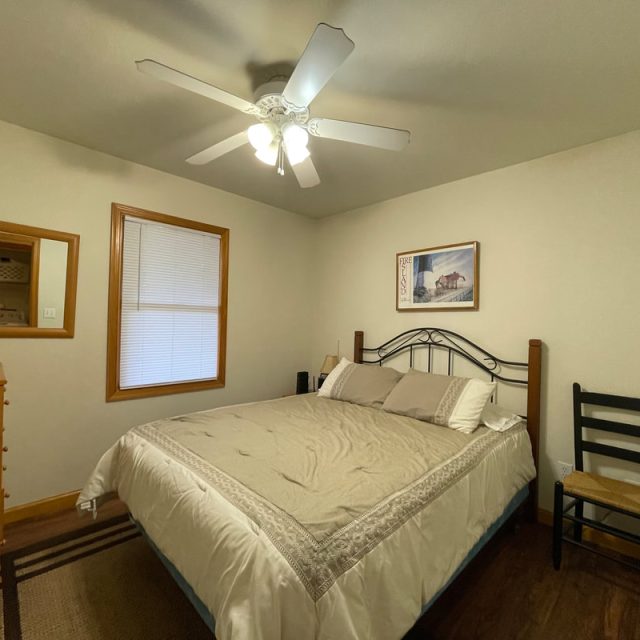 A small bedroom with a double bed, wooden dresser, TV, chair, ceiling fan, and framed wall art.