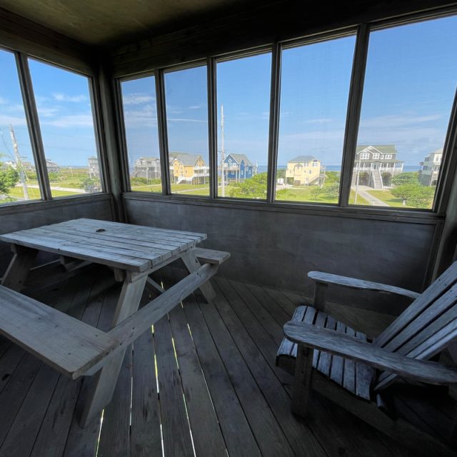 Screened porch with a wooden picnic table and chair, overlooking houses and a blue sky through large windows.