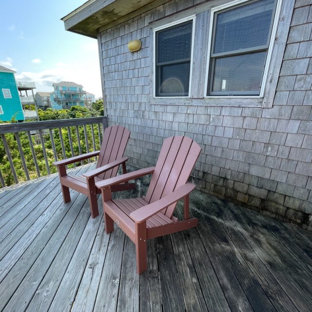 Two wooden chairs sit on a weathered deck outside a shingle-sided house, with other houses visible in the background.