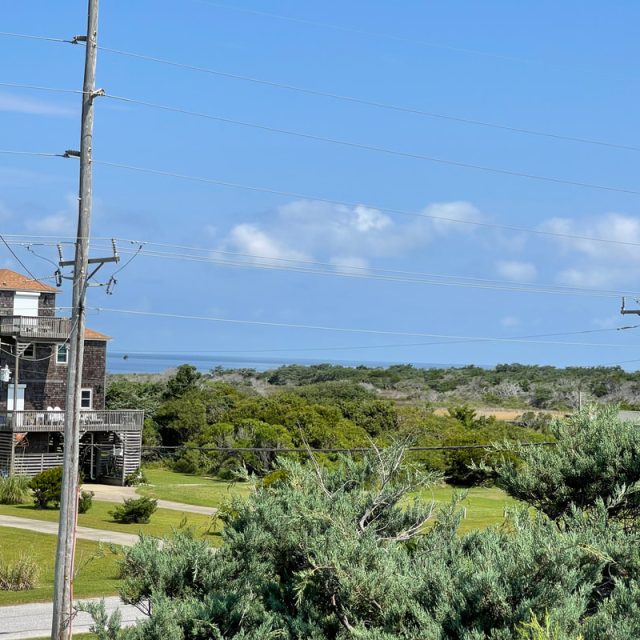 Wooden house near utility poles, green bushes, and a distant view of trees and ocean under a blue sky with clouds.