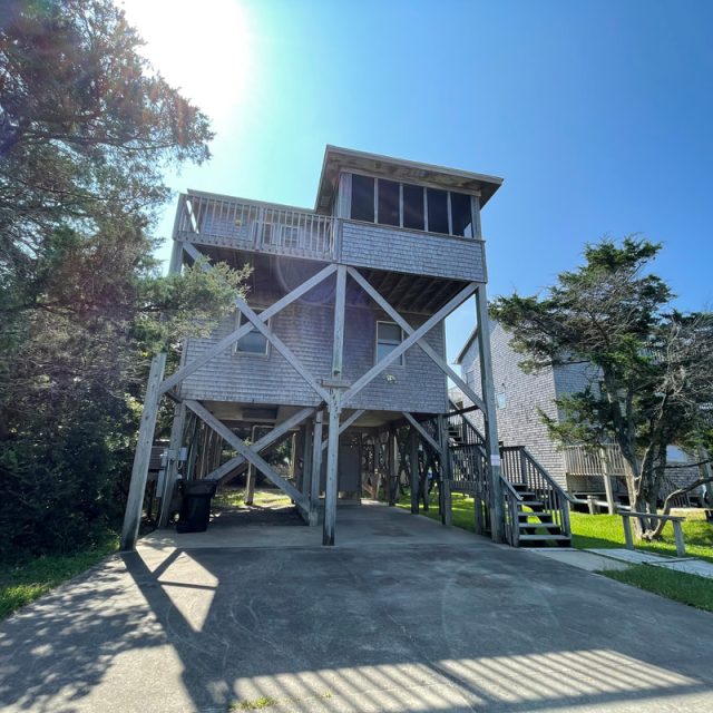 A stilted wooden beach house with a balcony, surrounded by trees on a clear sunny day.