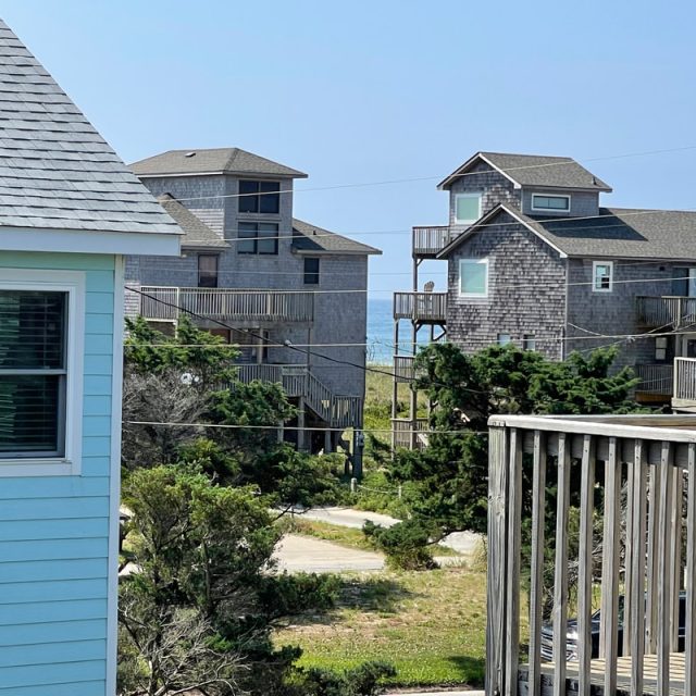 Several coastal houses with wooden decks and distant ocean view, framed by trees and part of a light blue building.